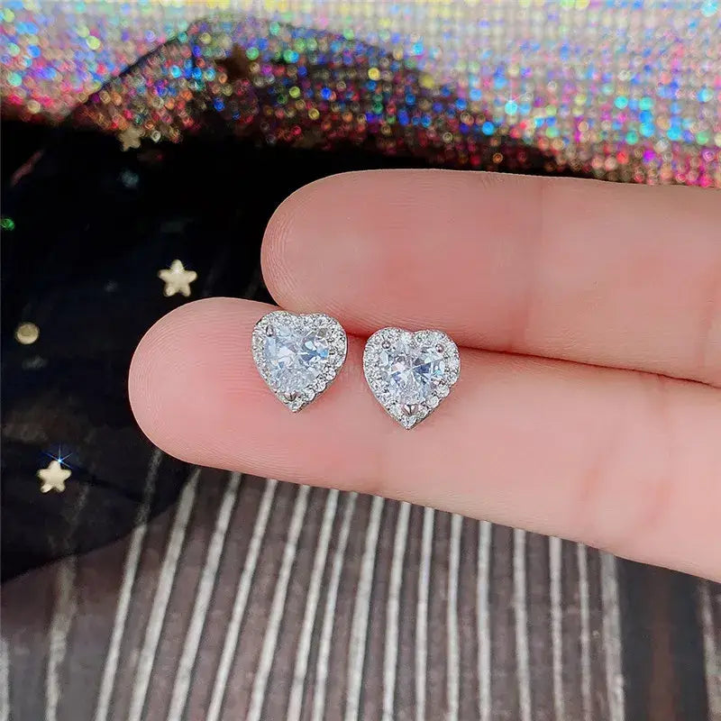 Heart-shaped diamond earrings held between fingers with a glittery background